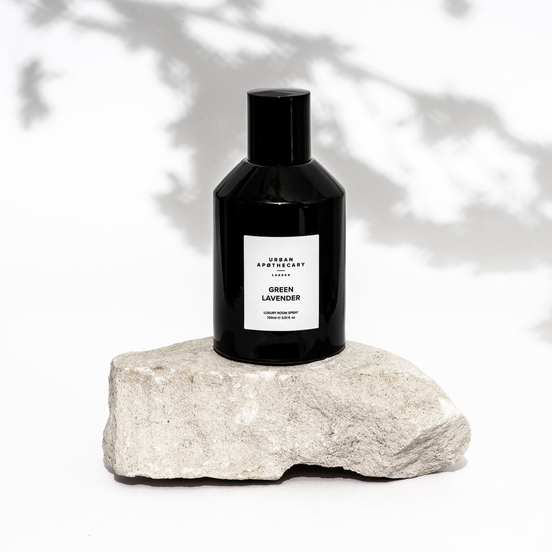 A black bottle of Urban Apothecary London Green Lavender room spray sits on a rough, light grey stone against a white background with soft leaf shadows.