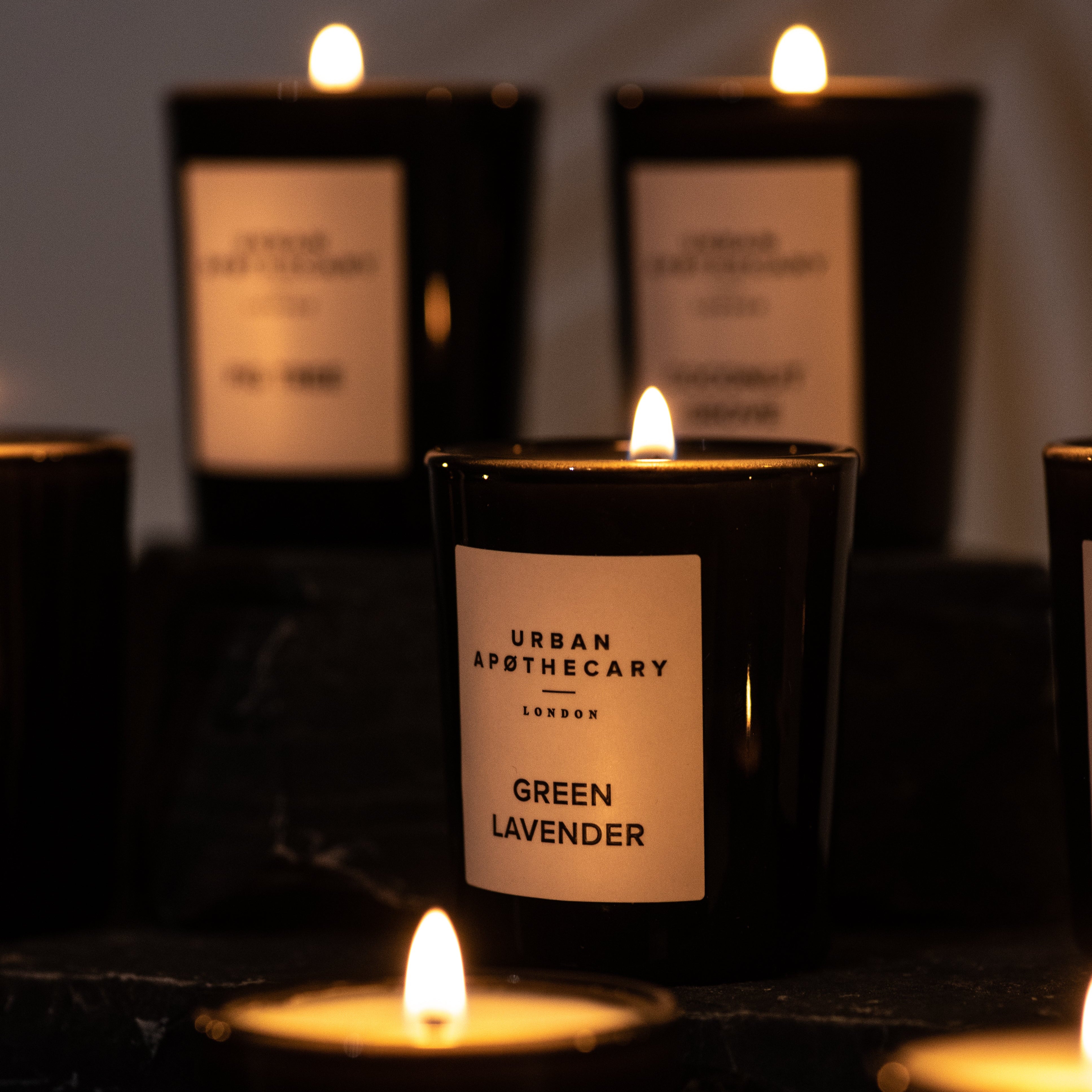 A lit black glass candle jar labeled 'URBAN APOTHECARY GREEN LAVENDER' in the foreground, with other matching candles blurred in the background on a dark surface.