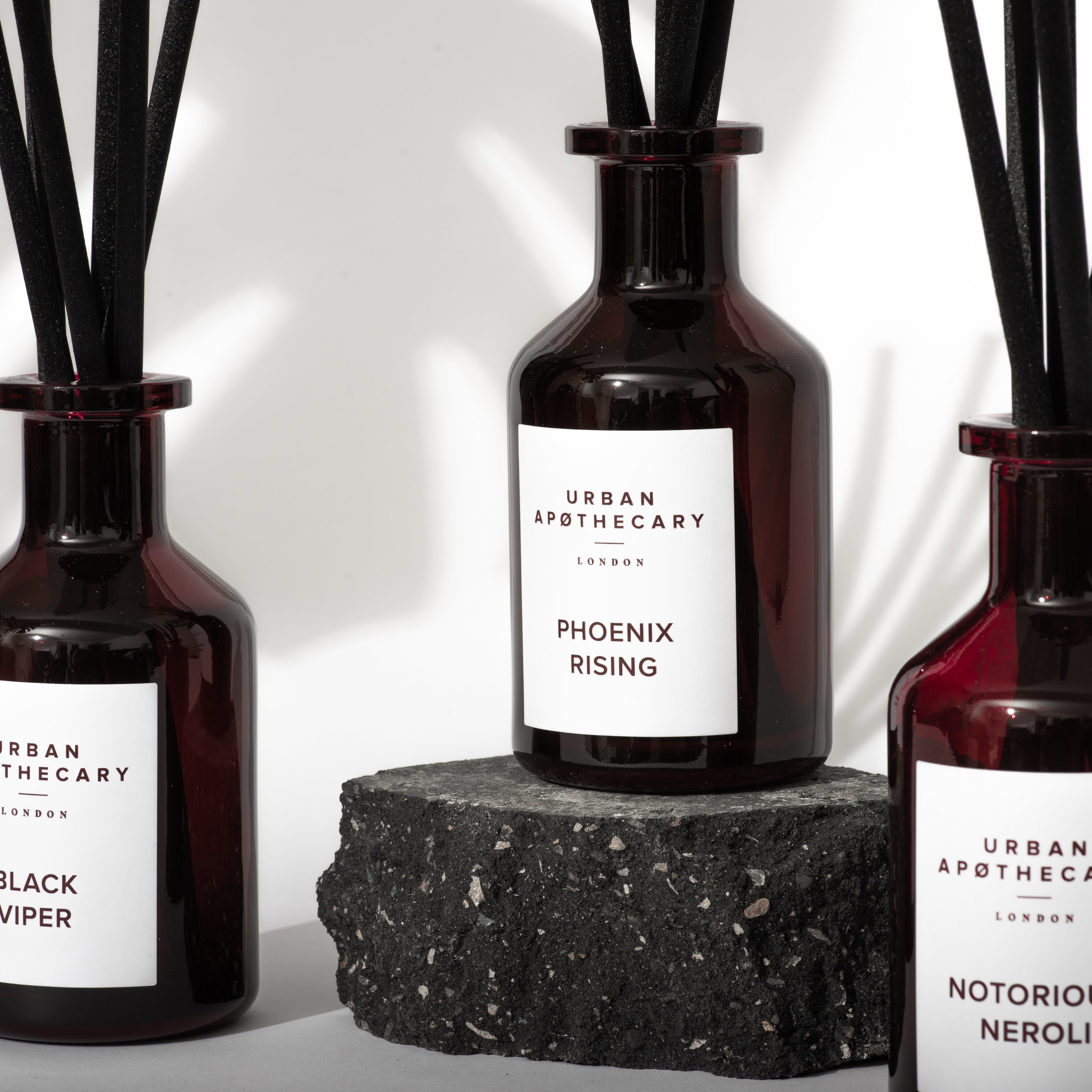 Three Urban Apothecary London reed diffusers in dark ruby glass bottles with black reeds, displayed on a white background with a textured stone block. The labels read Black Viper, Phoenix Rising, and Notorious Neroli.