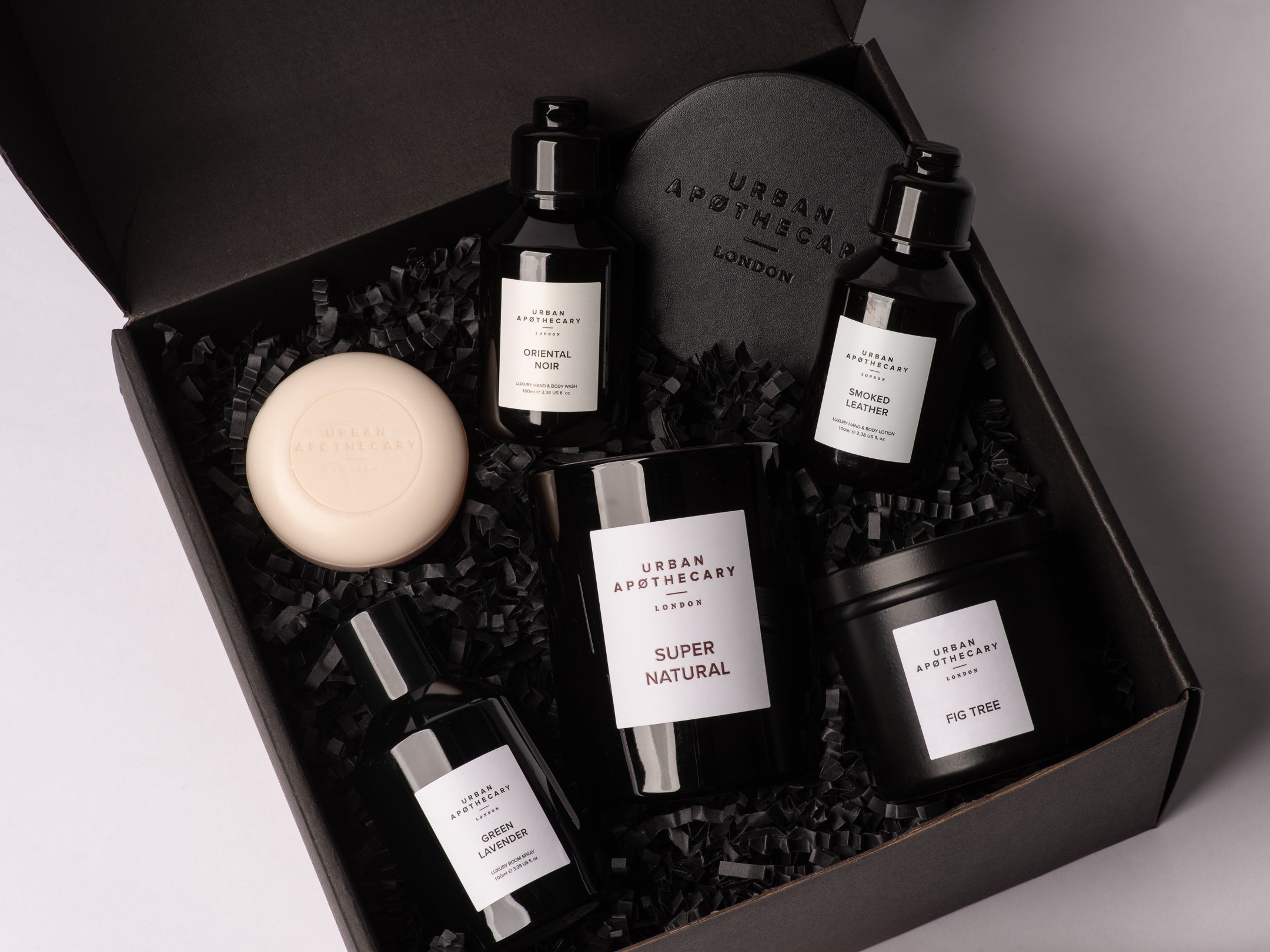 Urban Apothecary London Mystical Surprise Box in a black box, featuring a Super Natural candle, Fig Tree candle, Oriental Noir Hand & Body Wash, Smoked Leather Hand & Body Lotion and a round Cherry Blossom cream-colored soap bar nestled in black crinkle paper.