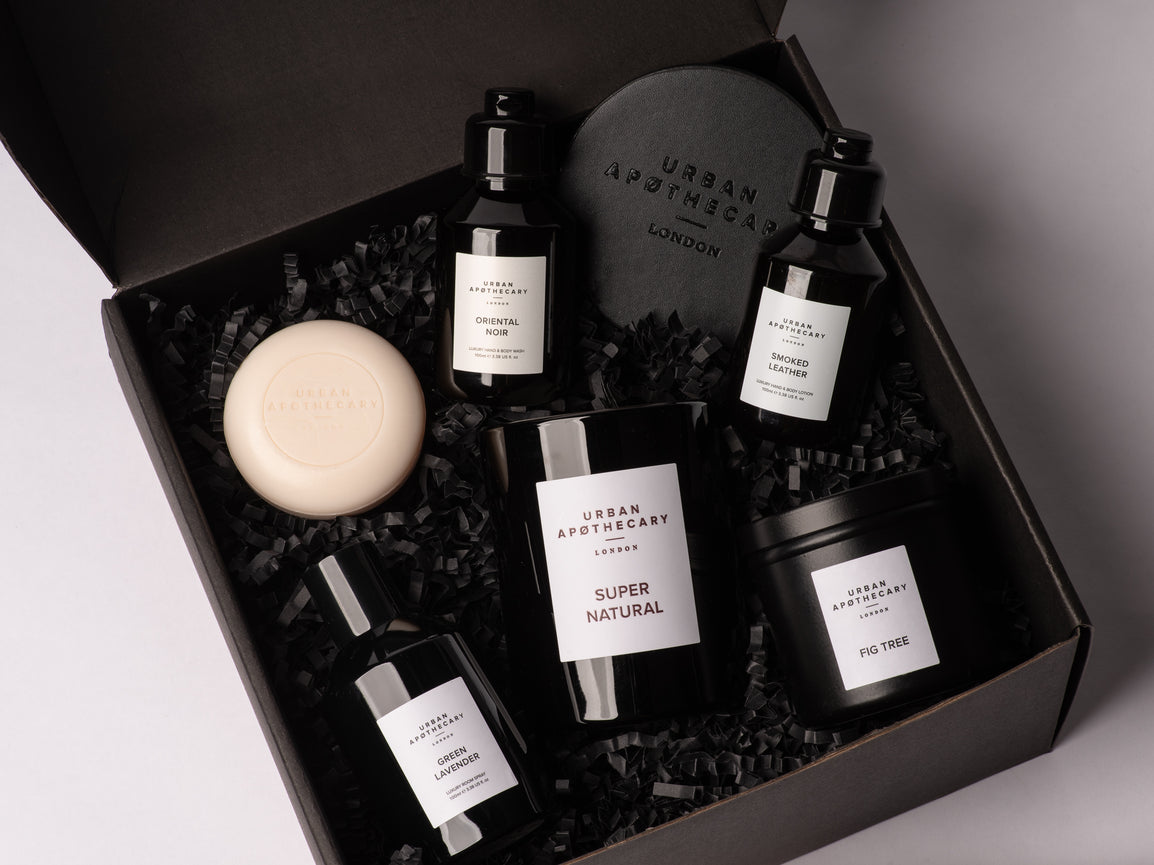 Urban Apothecary London Mystical Surprise Box in a black box, featuring a Super Natural candle, Fig Tree candle, Oriental Noir Hand & Body Wash, Smoked Leather Hand & Body Lotion and a round Cherry Blossom cream-colored soap bar nestled in black crinkle paper.