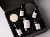 Urban Apothecary London Mystical Surprise Box in a black box, featuring a Super Natural candle, Fig Tree candle, Oriental Noir Hand & Body Wash, Smoked Leather Hand & Body Lotion and a round Cherry Blossom cream-colored soap bar nestled in black crinkle paper.